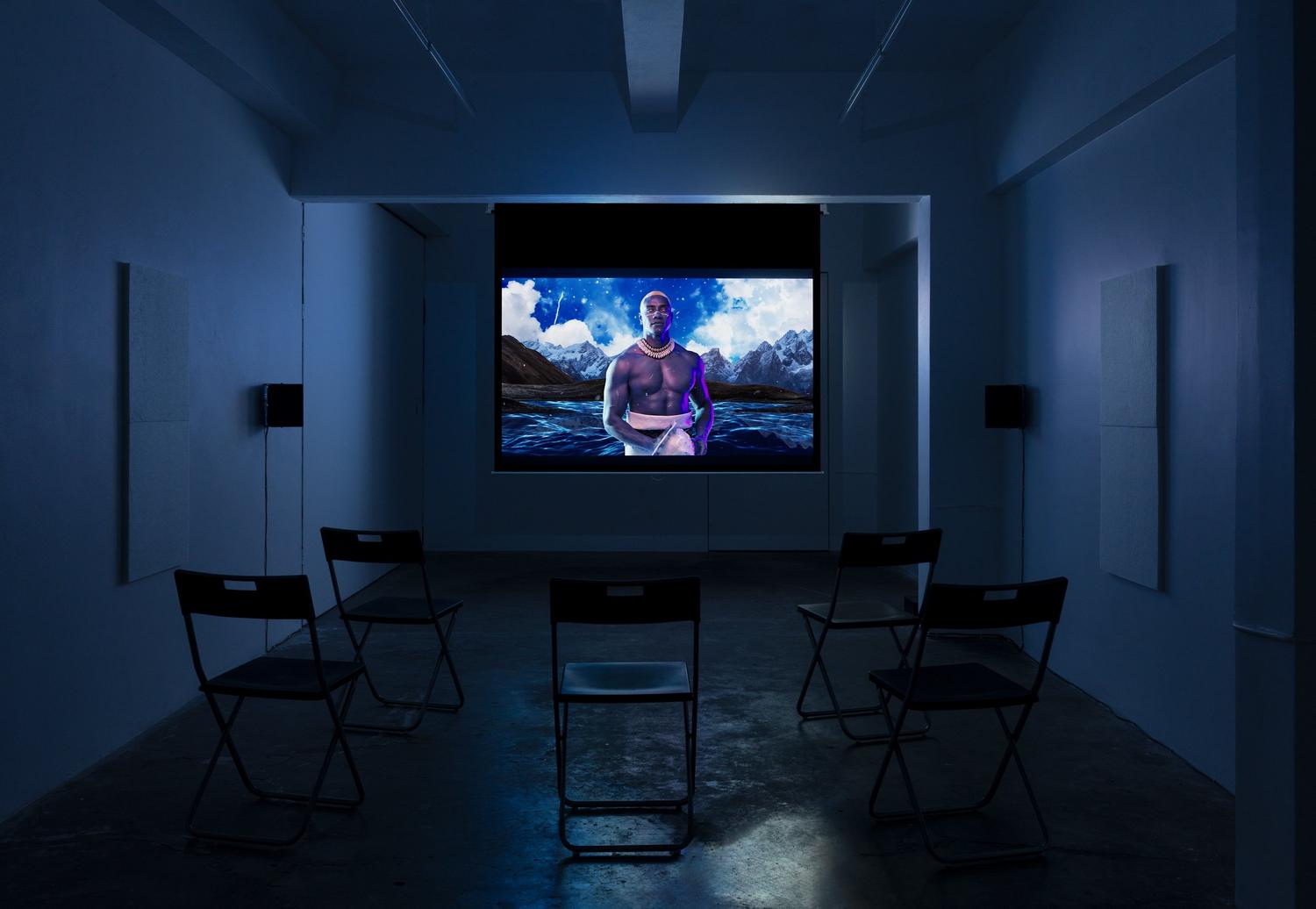 A screen play in a darkened room (blue hue): a man in traditional Samoan dress stands in front of a digital landscape of ocean and mountains.