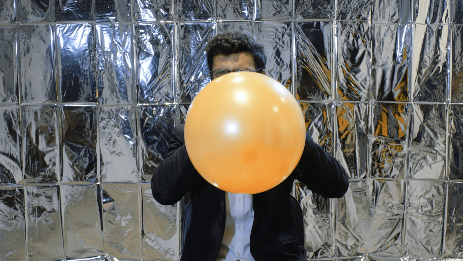 A person's face is obscured by a balloon which they are inflating close to bursting