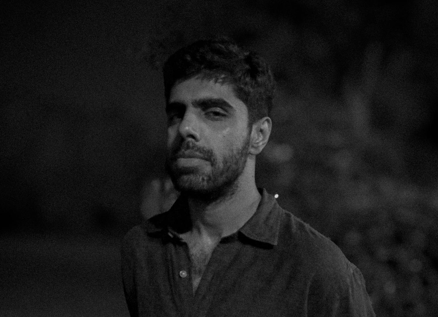 In dark moody light a person looks unexpressively at the camera