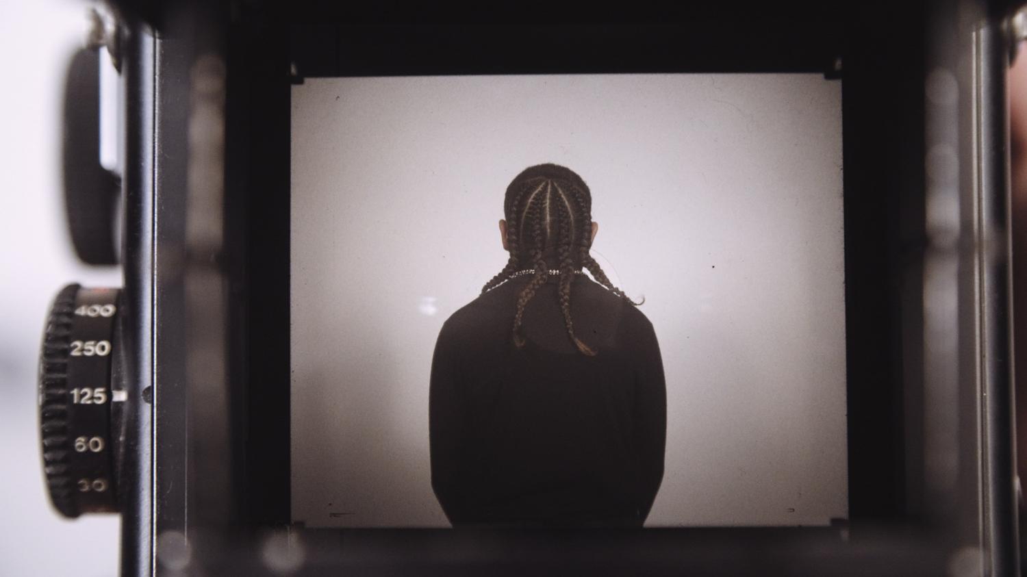 Seen through the viewfinder of a camera, we see an image of a youth with braided hair. They are dressed in black and stand with their back to camera. At left we see the dials of the camera apparatus, used for setting exposure, light level and other functions.