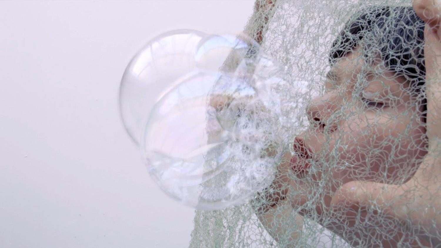 A woman blow bubbles througn a net. Her eyes are closed and her hands push the net to tension it as she blows through it.