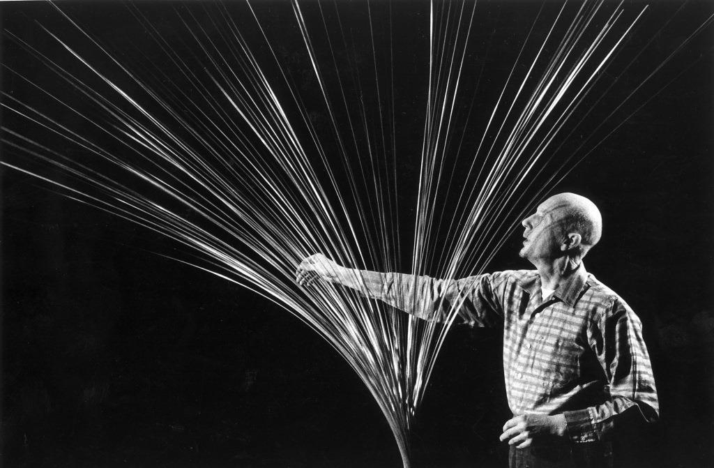 Artist Len Lye reaches towards a sculpture made of metal rods fanning out from the base. Lye's gaze is fixed as if to gauge the movement of the metal rods, and his had raches as if to steady one of the rods.