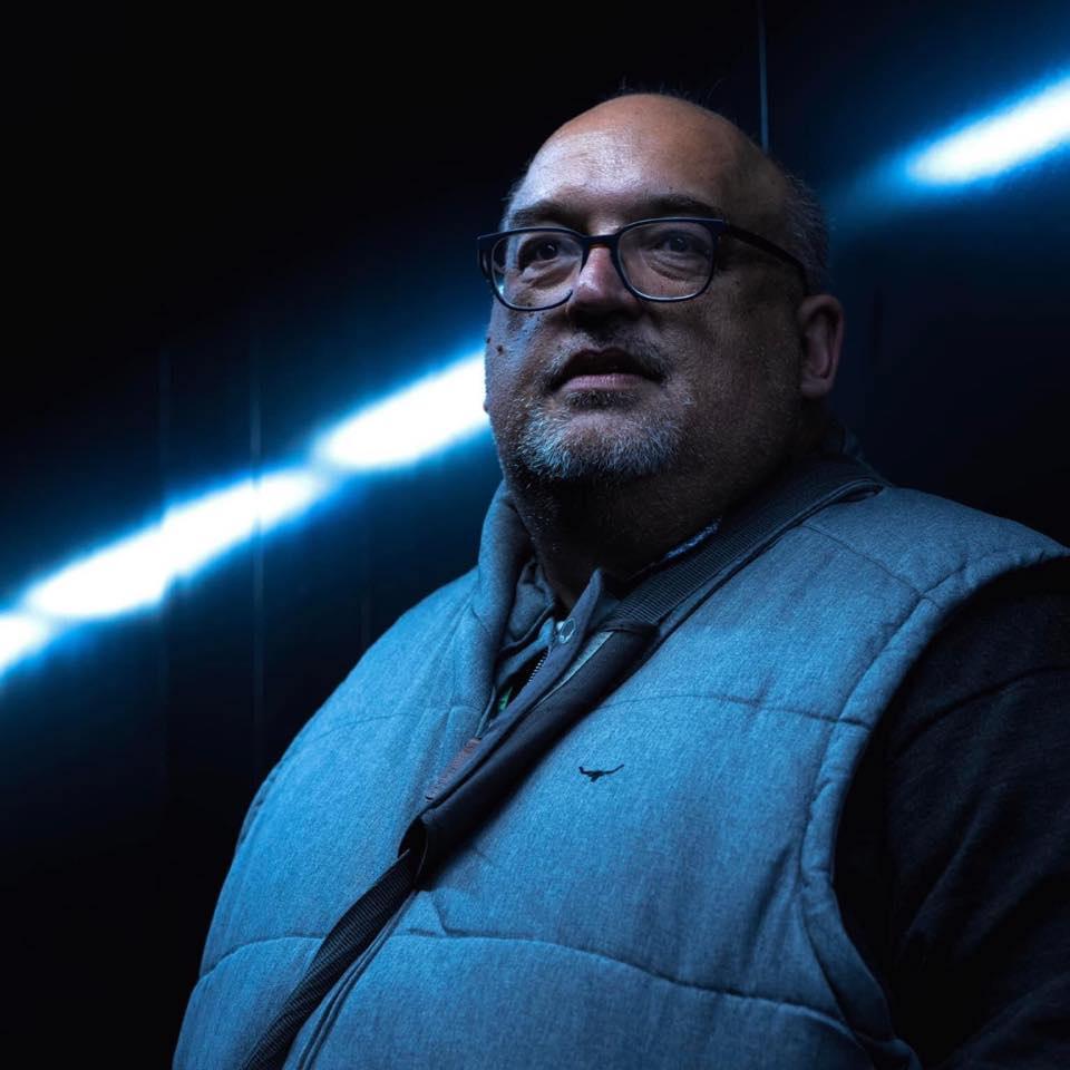 Image of a man with glasses and facial hair, wearing a puffer vest against a dark background