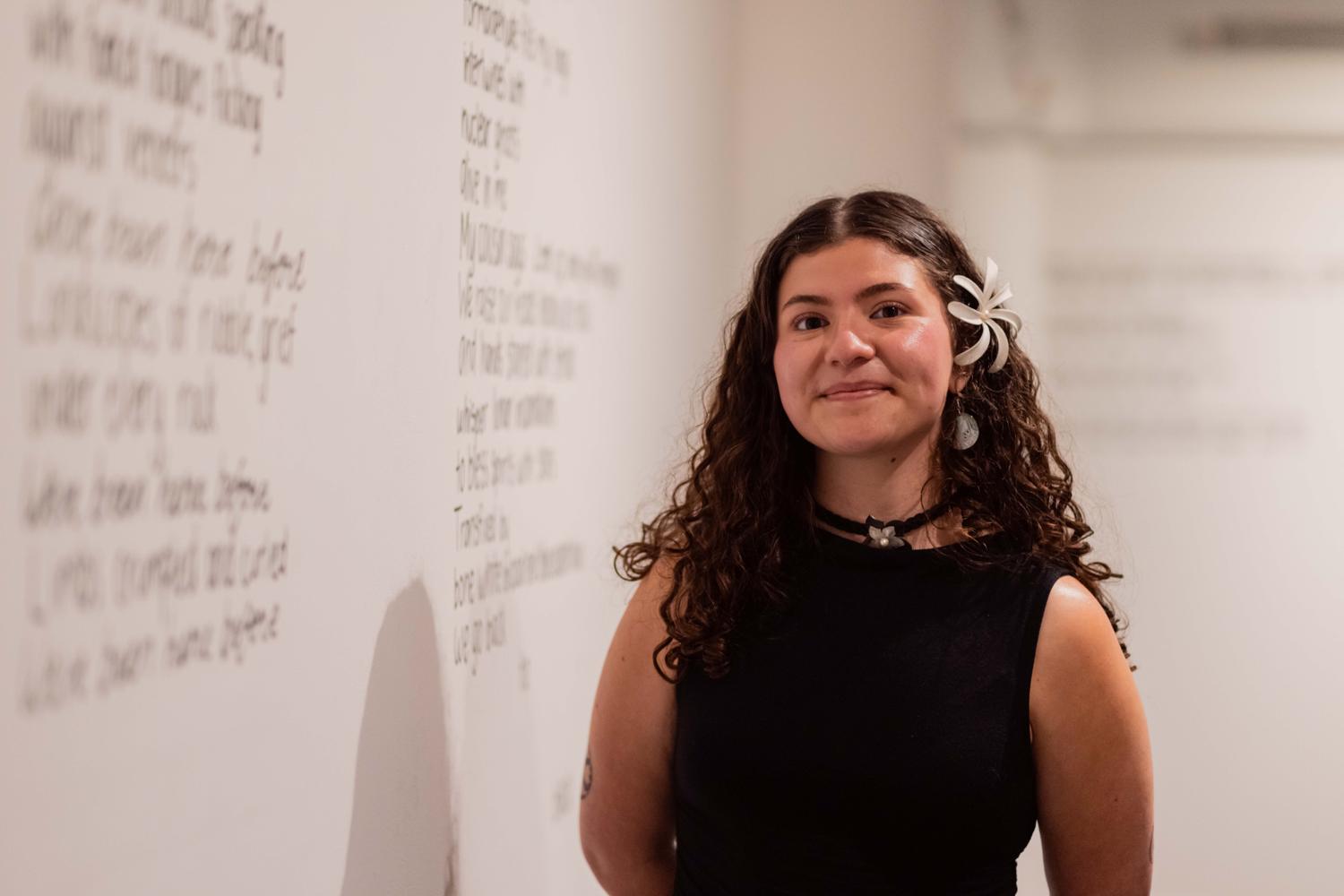 A young woman in semi-formal attire stands besdie a gallery wall. She is smiling and has a flower in her hair. On the wall is blurry text.