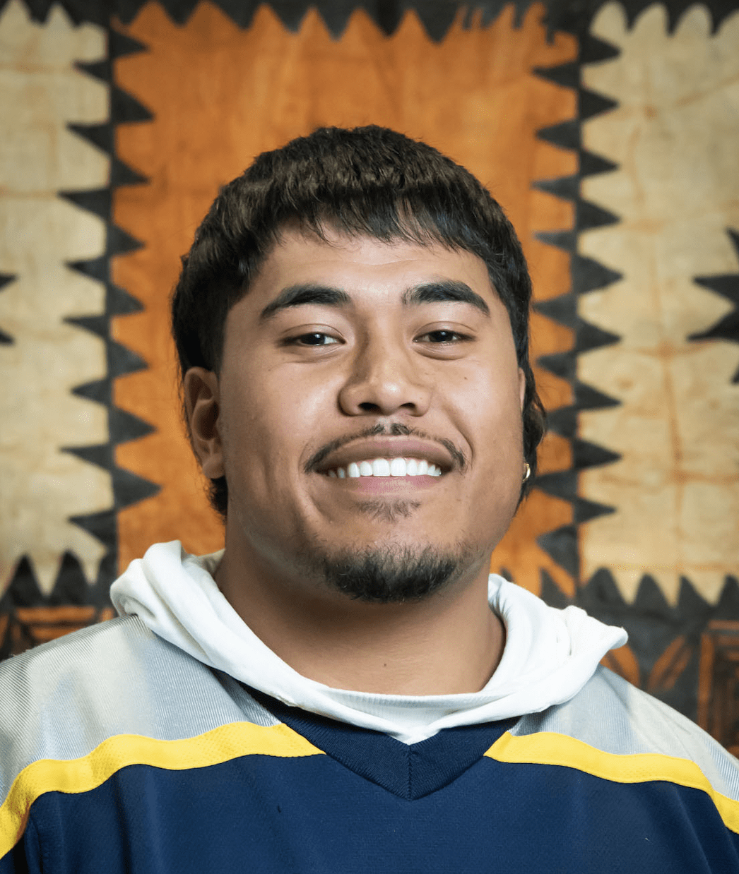 A man smiles at the camera; printed barkcloth or tapa can be seen in the background