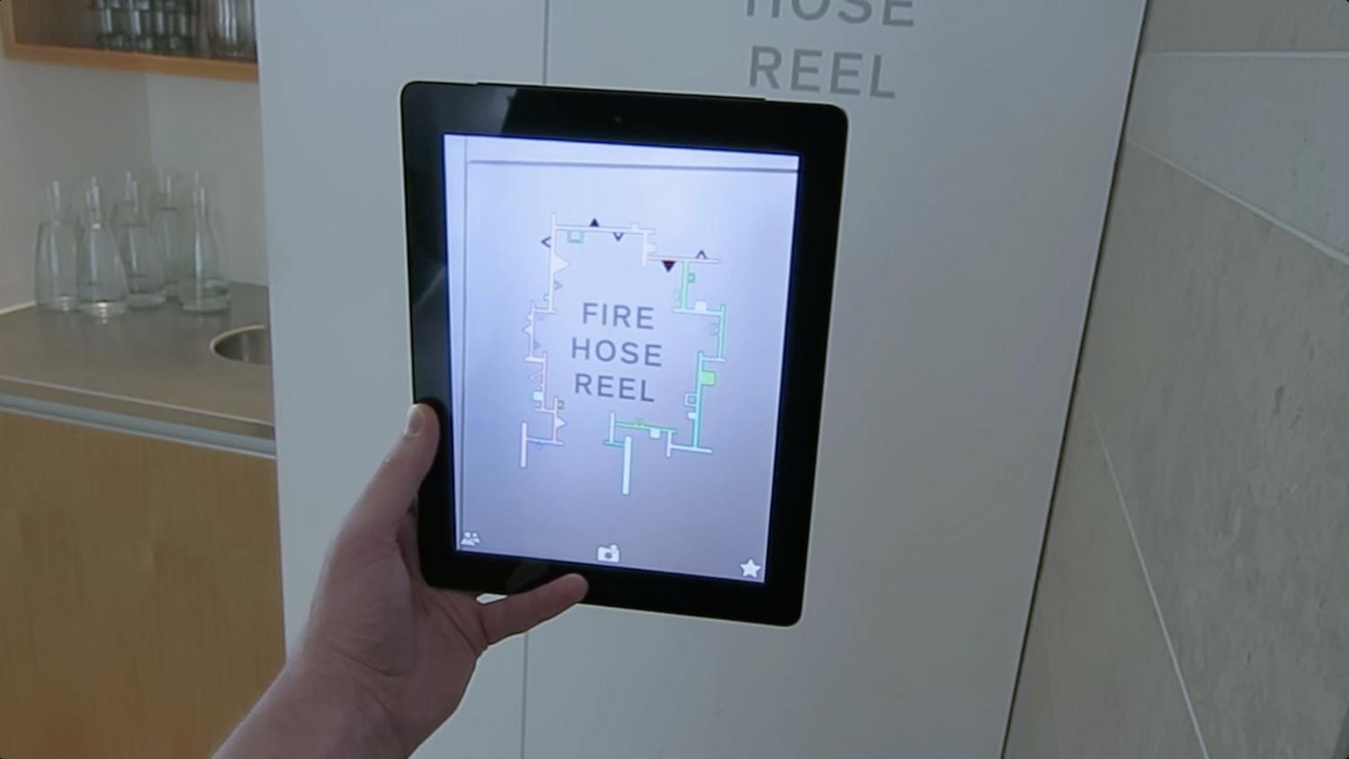 A computer tablet held in front of a sign shows the text 'Fire Hose Reel' augmented with a series of geometric shapes and details