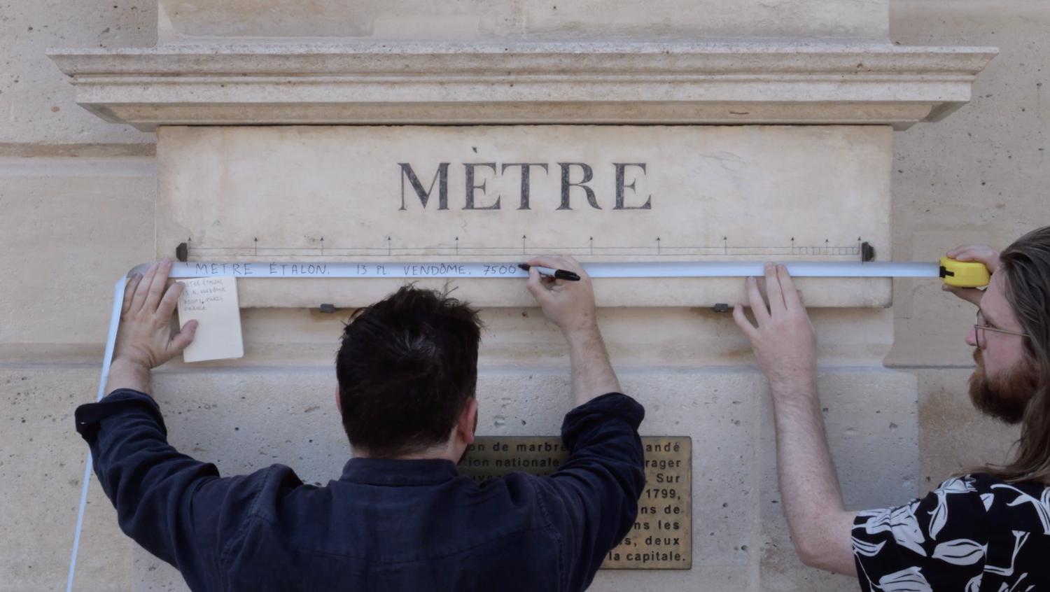 Two people use a measuring tape to gauge the length of a standard metre stone block.