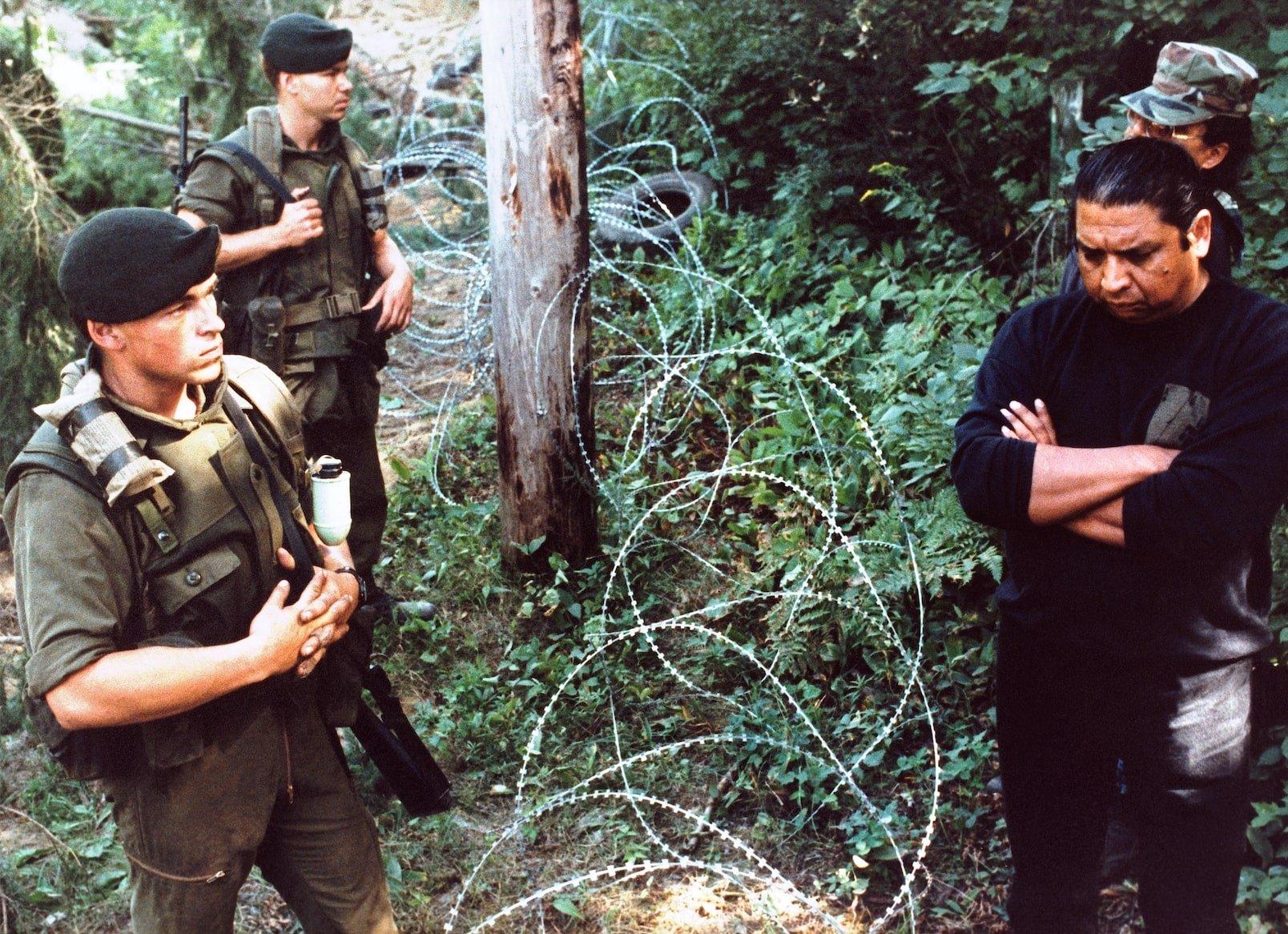 Two groups of men stand on either side of barbed wire in a forest. One side wears military uniforms.