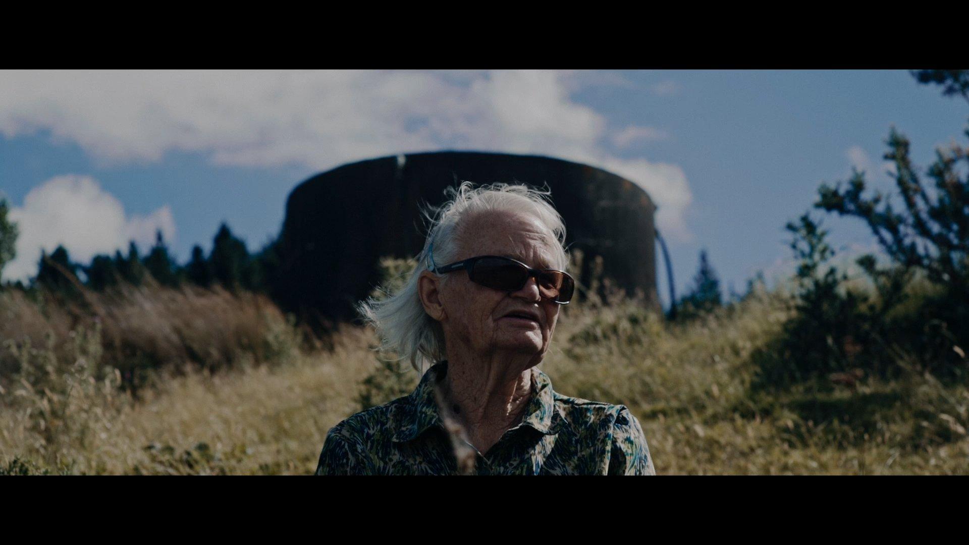 A older person wearing sunglasses stands downhill from a hilltop watertower. She appears to be speaking to a memory of the place.