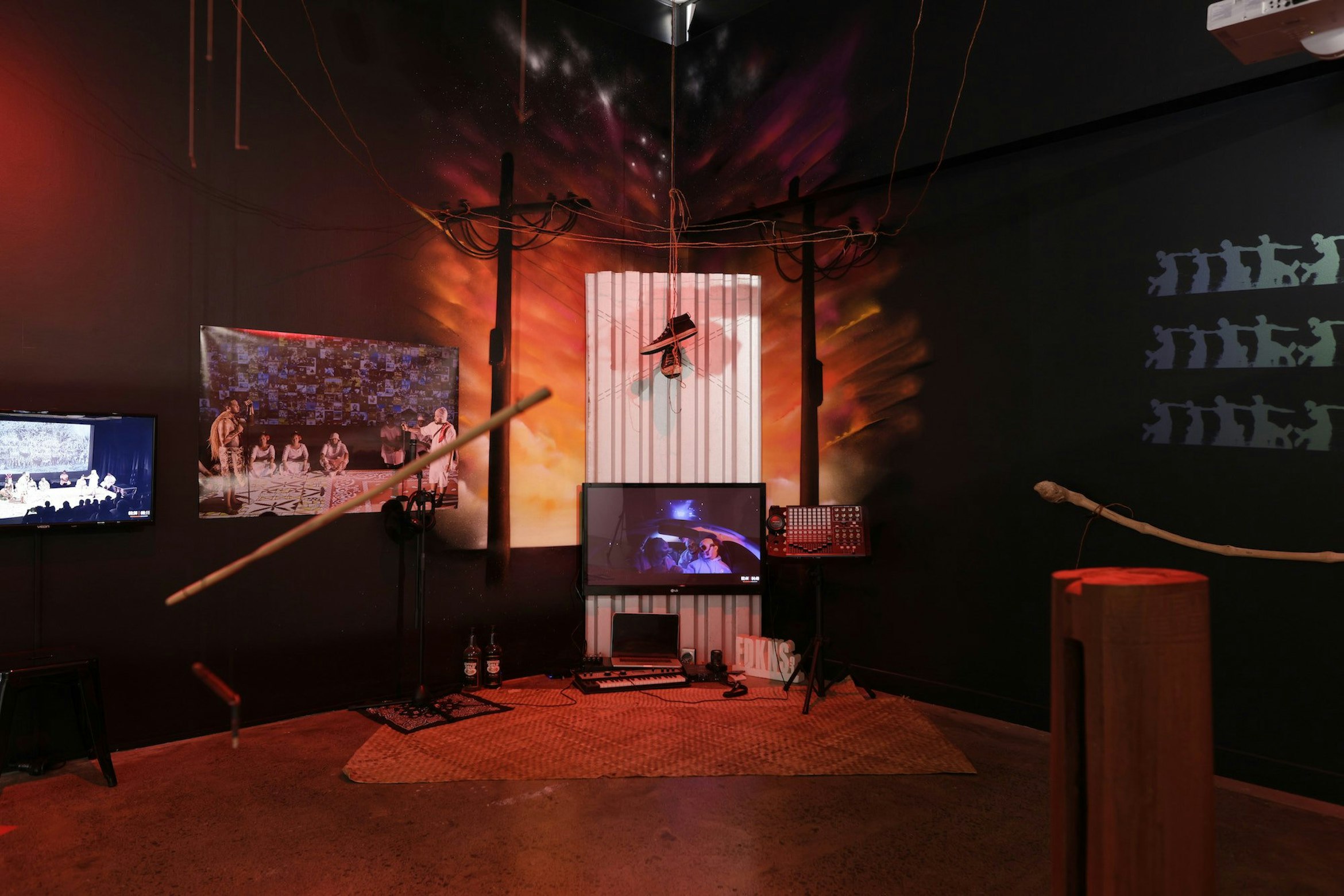 A dense multi-media installation including TV monitors, a pair of hanging sneakers, a computer, Pasifika musical instruments, stencil on the wall and more.