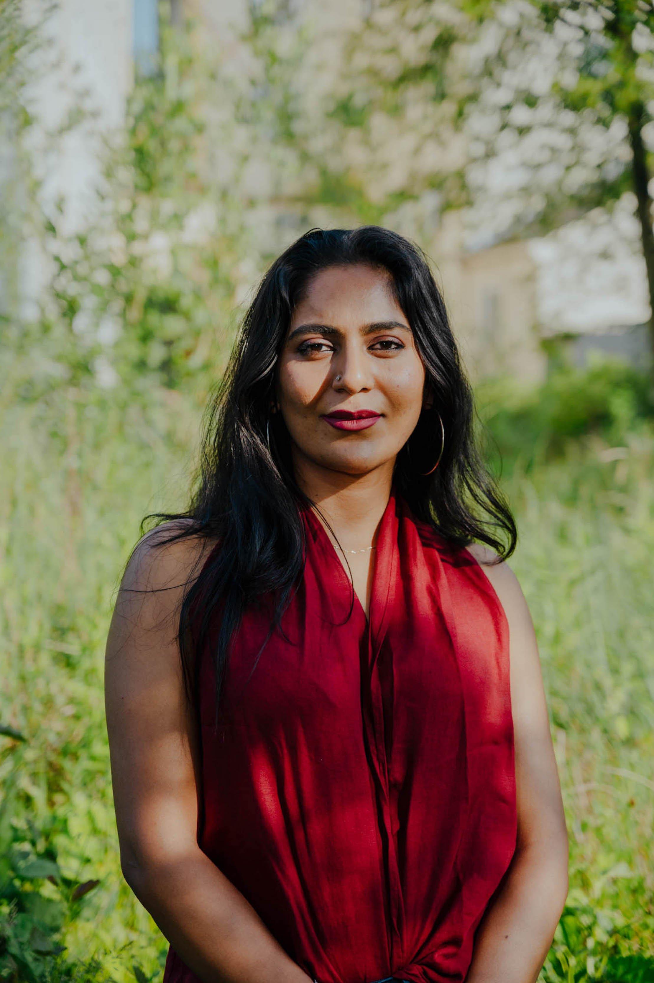 A woman in a red dress stands outside in front of greenery and looks straight at the camera.