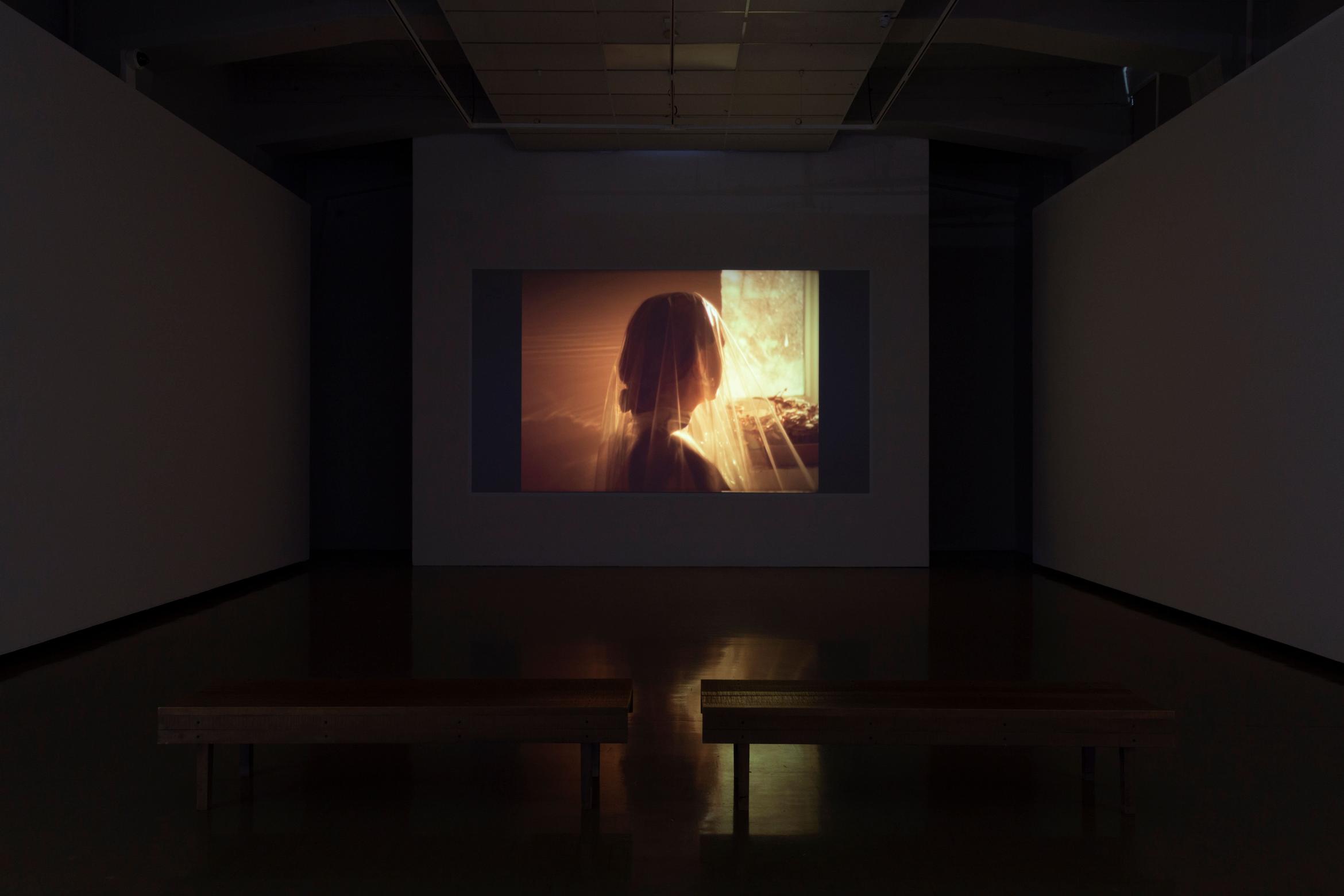 A woman in a sheer veil faces away from the camera; the light is golden. An installation view of a screen in a darkened room.