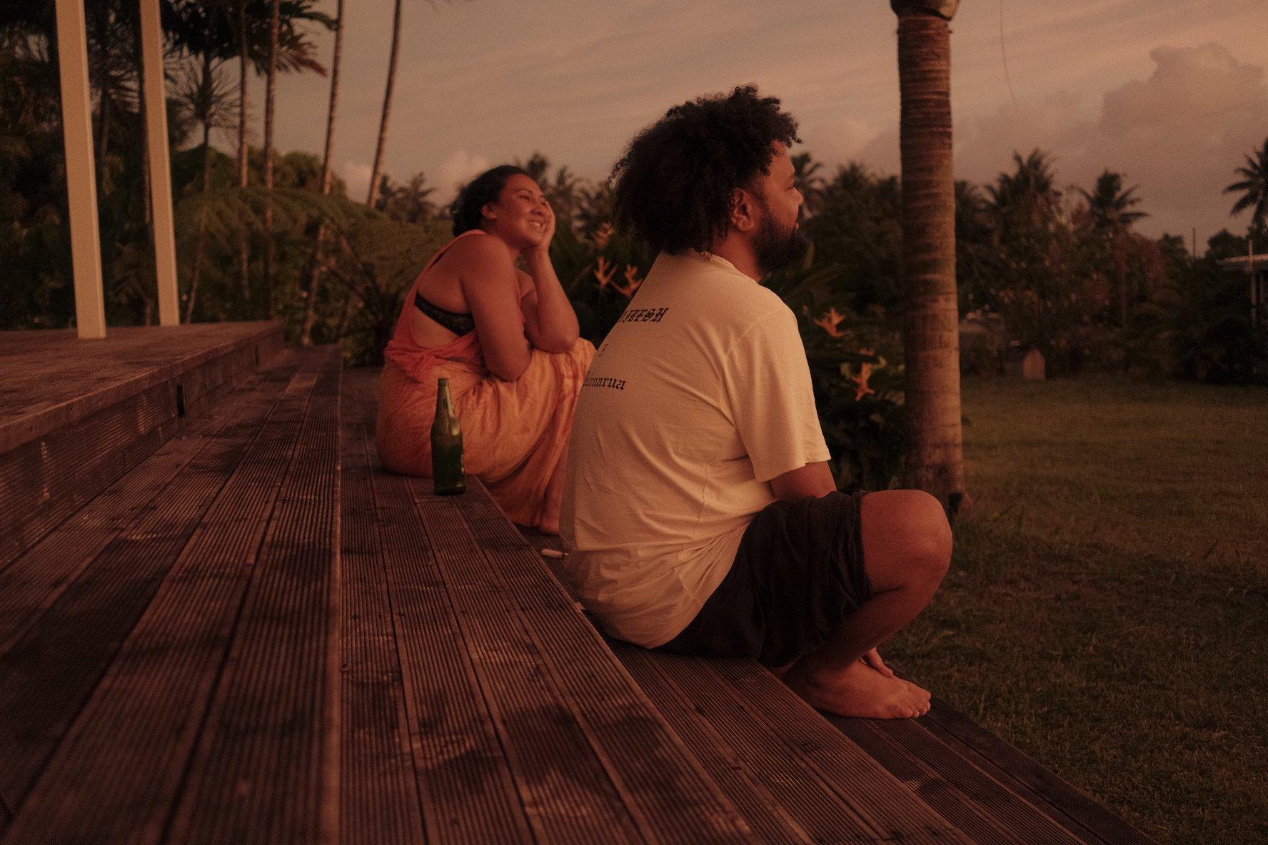 A man and a woman sit on the edge of a deck with beers in the last light of the day, the setting is in Samoa