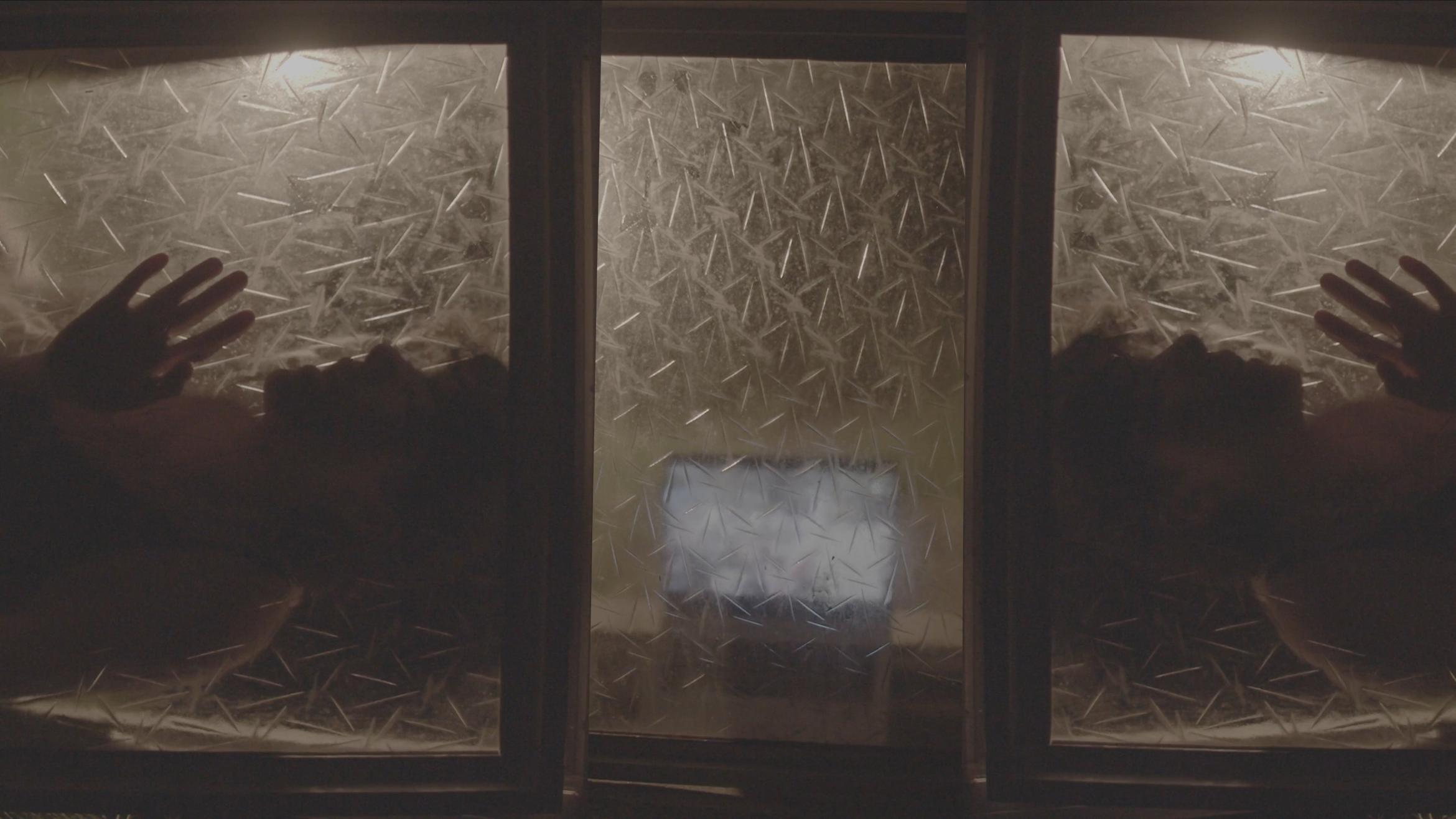 Three frosted windows are lit up from behind by lamps. One on each side reveal the silhouette of someone caressing the window, while the middle window shows a laptop playing a video.