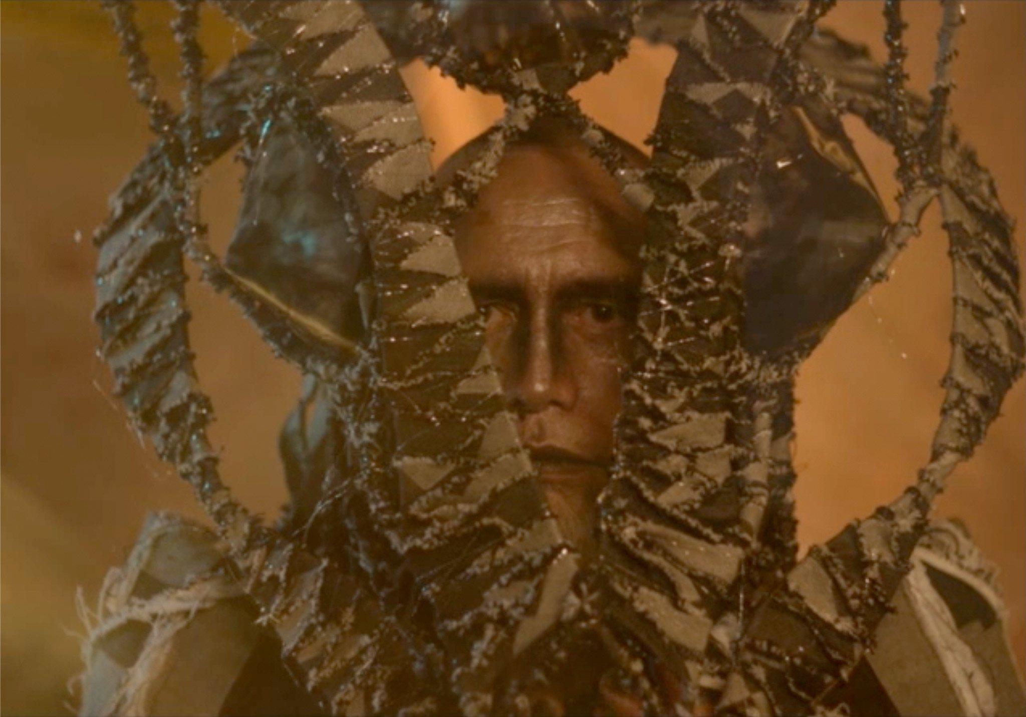 The face of a male figure shows a stern expression. Their head is enveloped by a series of green wavy tendrils, which point upright.