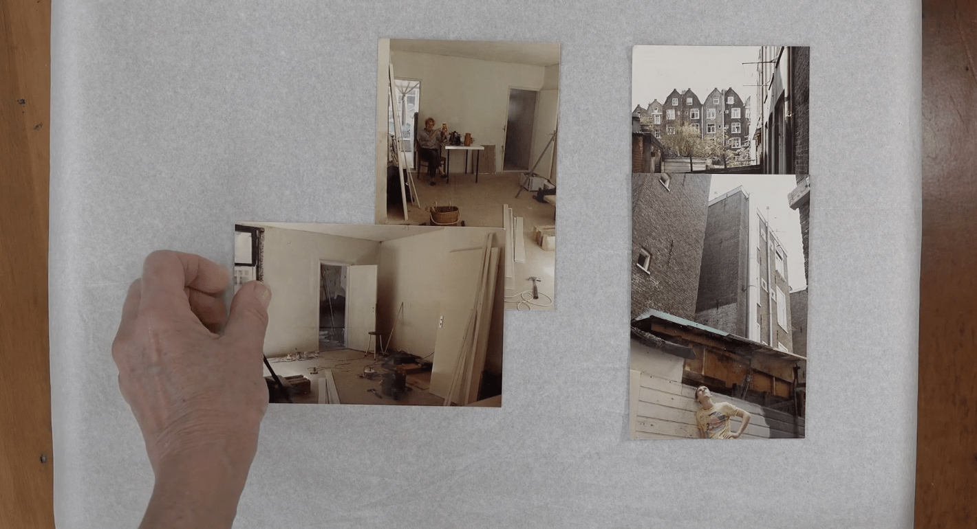A hand arranges photographs of a house under construction on a large white sheet of paper