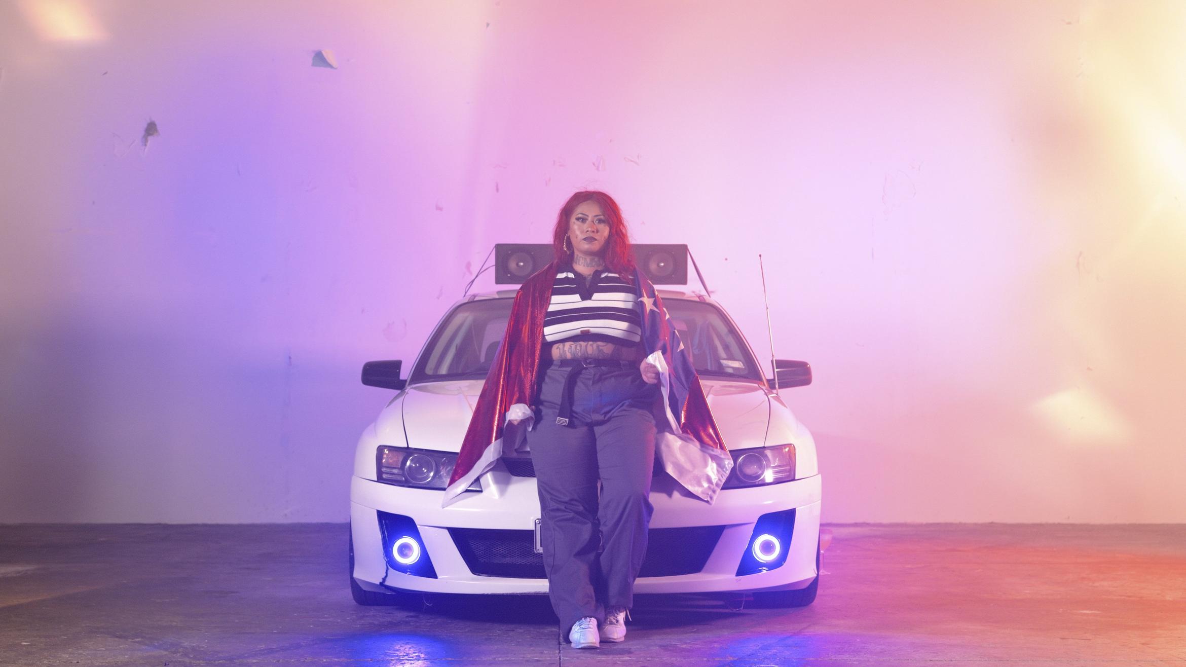 A young Pasifika woman leans steadfastly against the bonnet of a boy racer style car, which has speakers mounted on the roof