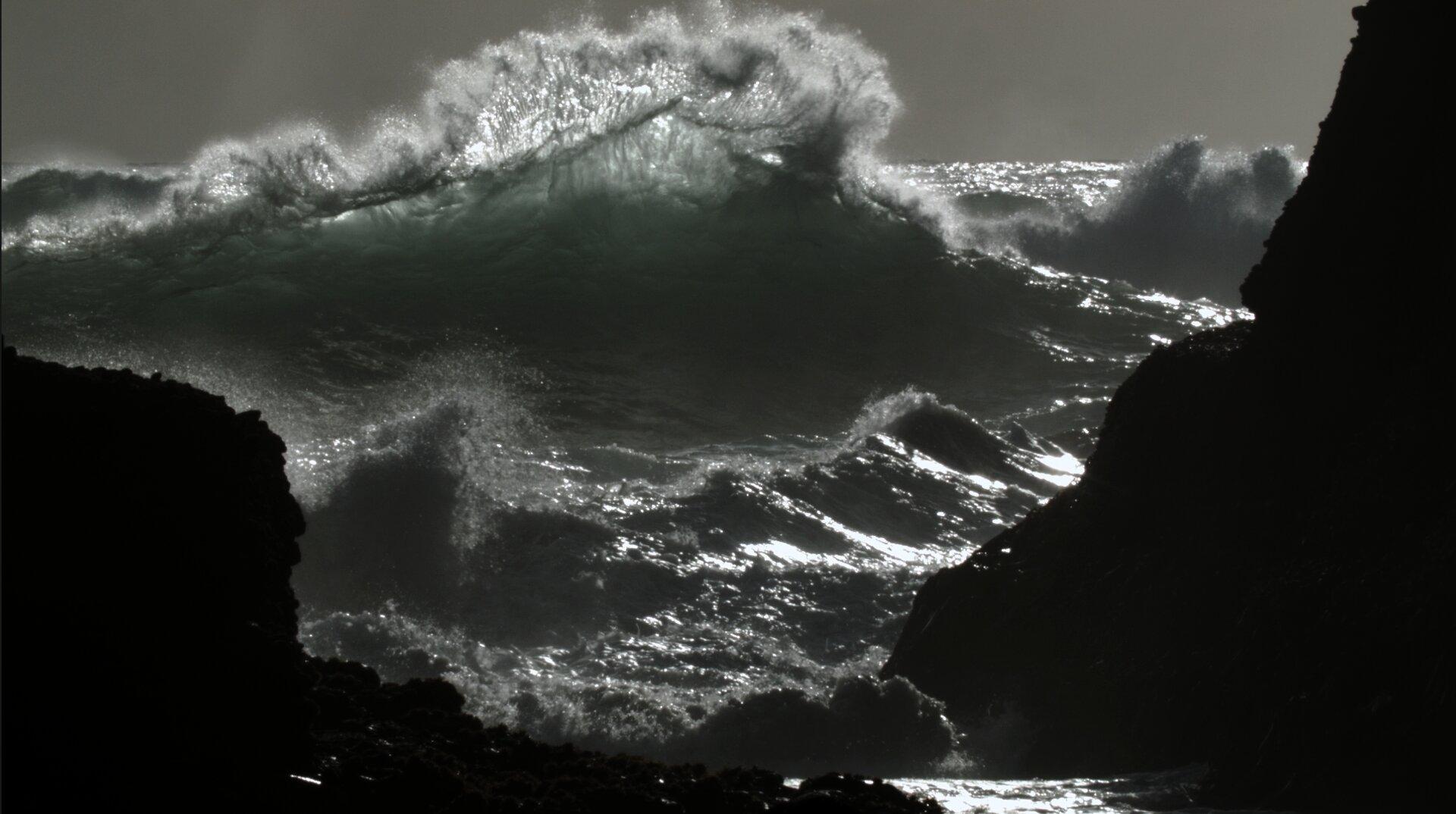 A stormy sea with a large wave breaking in the middle and rocks seen close by.