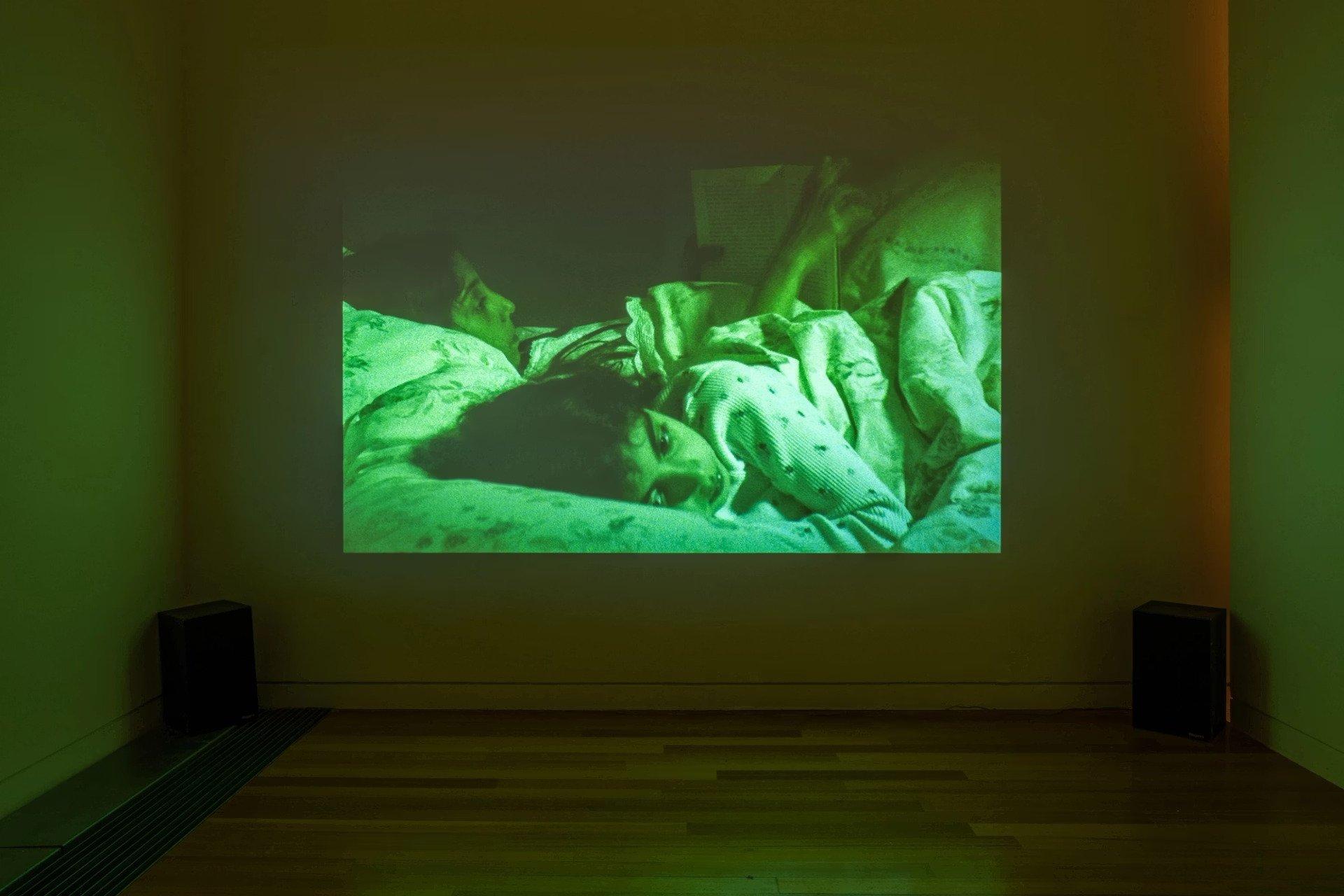 In a gallery we see a projected image of two young people in bed, each of whom is awake at night. The image is cast in a green luminescent hue.
