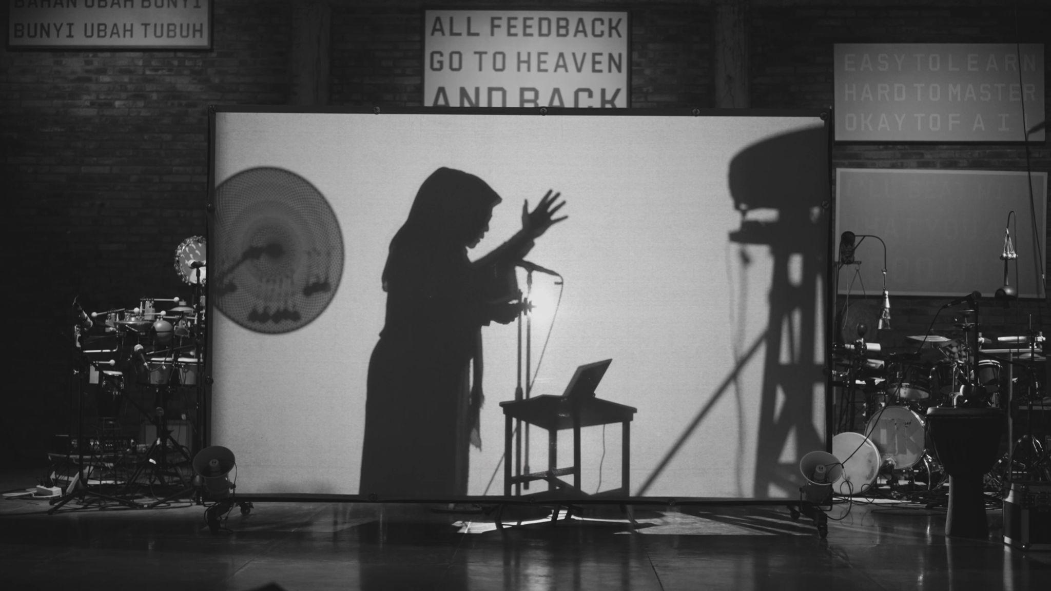 Behind a screen we see the shadow of a woman gesturing upwards whilst speaking into a microphone. At either side of the screen are drums and percussion. On the wall are texts in Indonesian and English. Above the screen reads "ALL FEEDBACK GO TO HEAVEN AND BACK".