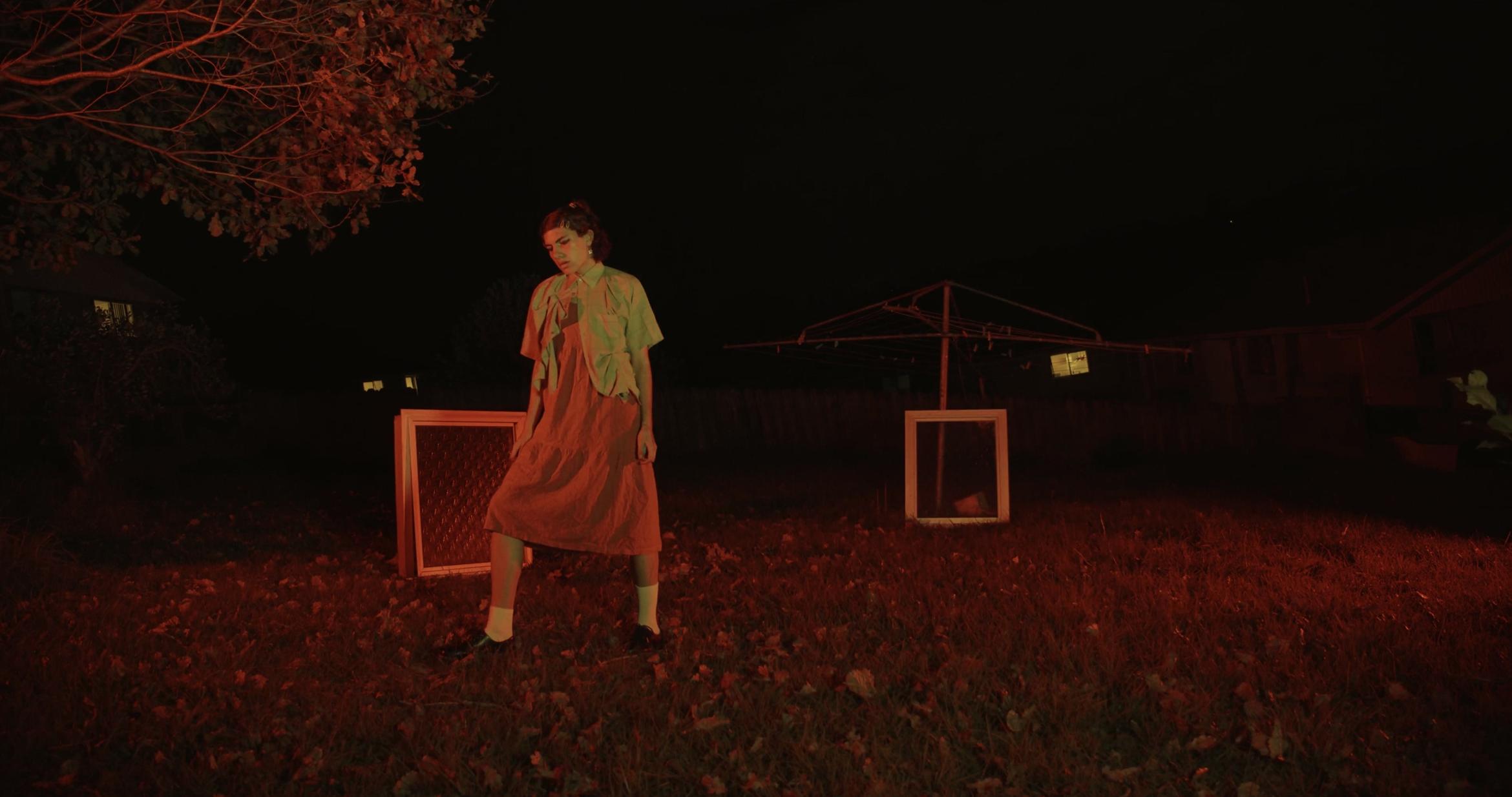 In a backyard a person in a dress stands in front of two windows which are propped up on the lawn. The second window rests against a clothes line. The scene is lit by a red glow.