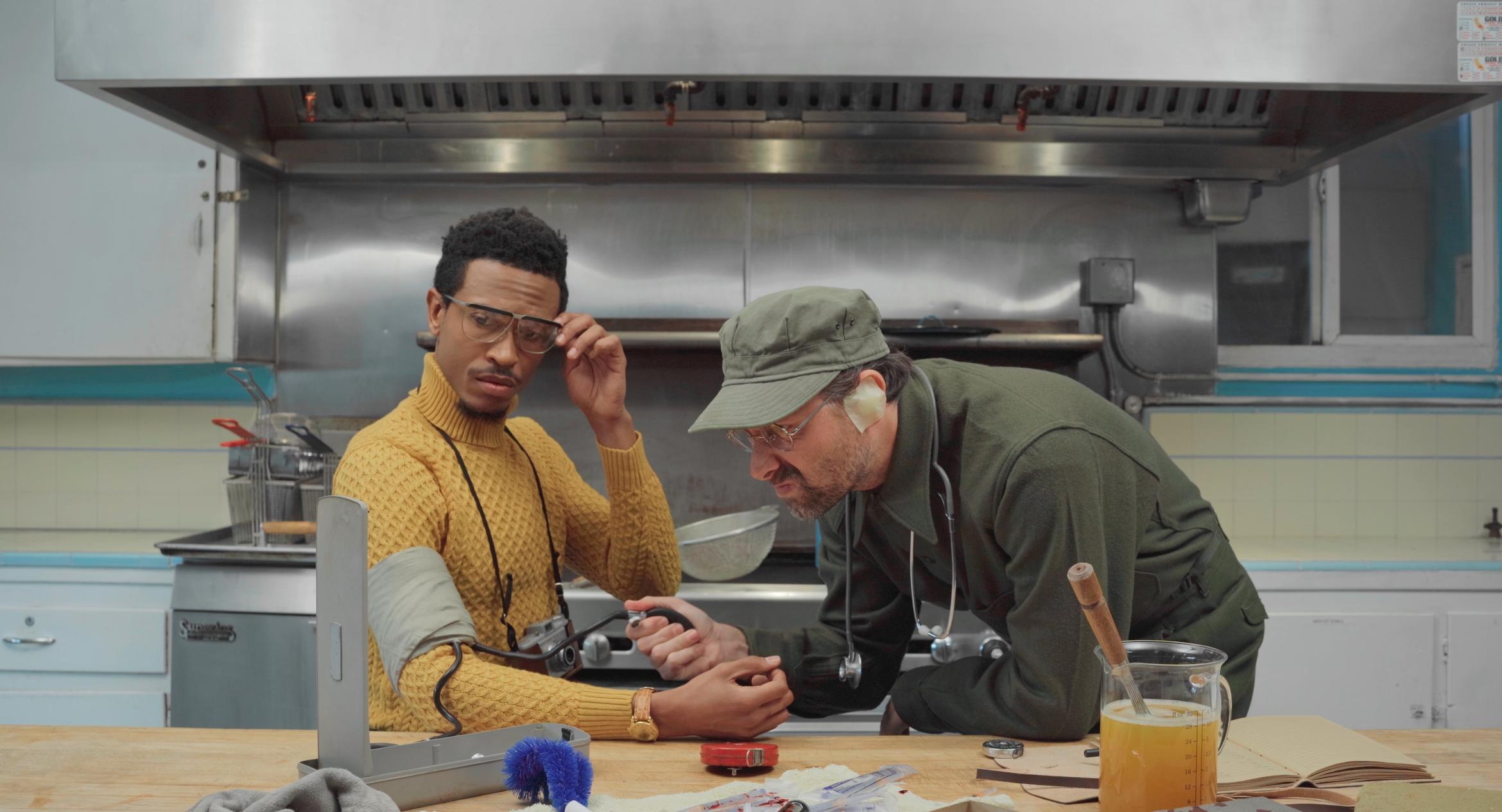 Two men in a kitchen perform vaguely medical or scientific actions