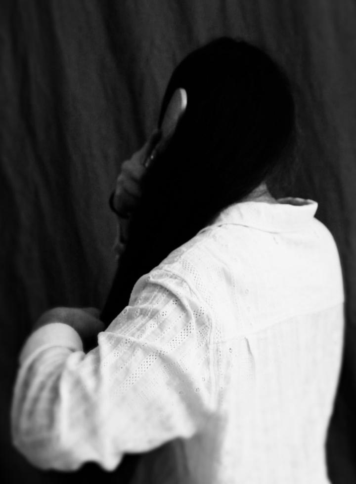 A black and white portrait of a person brushing their hair. They face away from the camera, their top is a white linen short or blouse. The image suggests a portraiture tradition from the Colonial era.