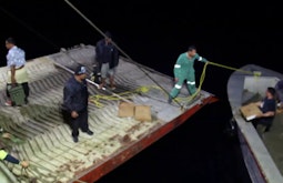 A group of people help bring a small boat closer to the back of a large ship to unload goods.