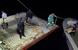 A group of people help bring a small boat closer to the back of a large ship to unload goods.