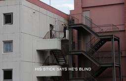 Two people stand on the stairs outside adjacent industrial buildings. Like the buildings the people are dressed in a monochromatic palette. Onscreen text says 'HIS STICK SAYS HE'S BLIND'.