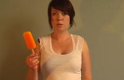 A person holds an orange popsicle up while looking into the camera.