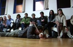 A person lies on the ground with a toilet plunger as a row of people look on.