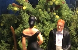 Two figurines of a man and a woman in wedding attire stand in front of a model tree.