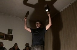 A person stands with a large shadow behind them as they bang two pots together.