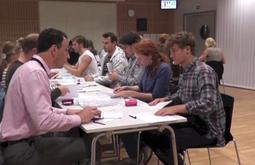 Participants in a performance sit at a long table in a conference room folding paper