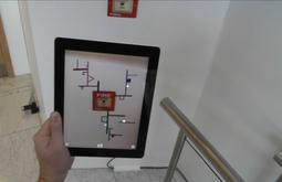 A person looks at a fire alarm switch through the lens of an iPad. Lines and geometric shapes are digitally drawn out from the switch.