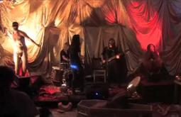 A band performs in a red and gold lit room, a naked man throws something from the corner.