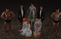 On a beach at night there are several people, two body builders, a man in a suit, and 2 women in long dresses.