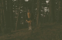 In a dark forest a person wearing a cheer-leader style outfit and boots does a dance to the camera.