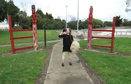 A child walks near a skate park. Two carved Māori figures are on either side of her.