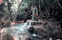 A person wearing a long white veil is superimposed onto a small waterfall in a forest.