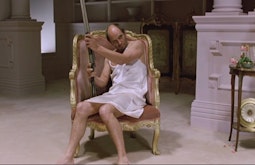A man sits in a gold gilded chair in a white slip dress holding a shotgun.