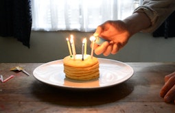 A hand reaches in lighting candles that are stuck in a stack of pancakes.