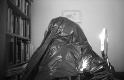 A person disguised in a rubbish bag holds a make up mirror to their face.