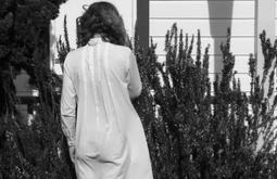 A person stands facing a rosemary bush by a house in a white dress.