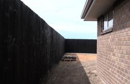 A brick house is surrounded by a tall black wooden fence, the ground is dry and bare.