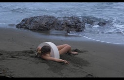 A person lies on sand by the waters edge, there is a semicircular sculpture over their torso.