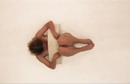 A birds-eye view of a person lying on a white floor with a semicircular sculpture across their torso.