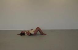 A person lies down in an empty white room with a bag of salt over their naked body.