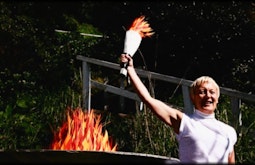 A person stands holding a victory torch made from paper beside a fire pit in which the fire is also made of paper.